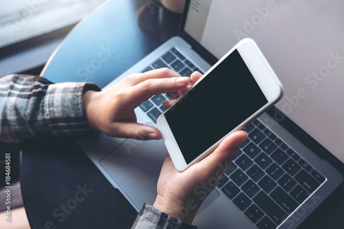 Mockup image of business woman holding mobile phone with blank black screen while using laptop on table in office