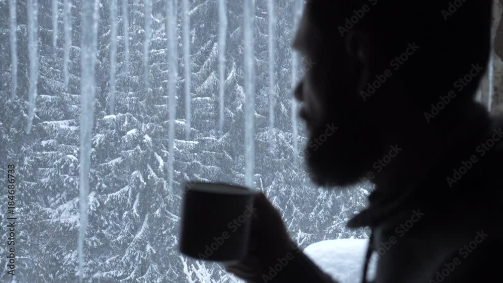 Handsome man drinking from a mug looking out of the window from inside house, wearing a black shirt.