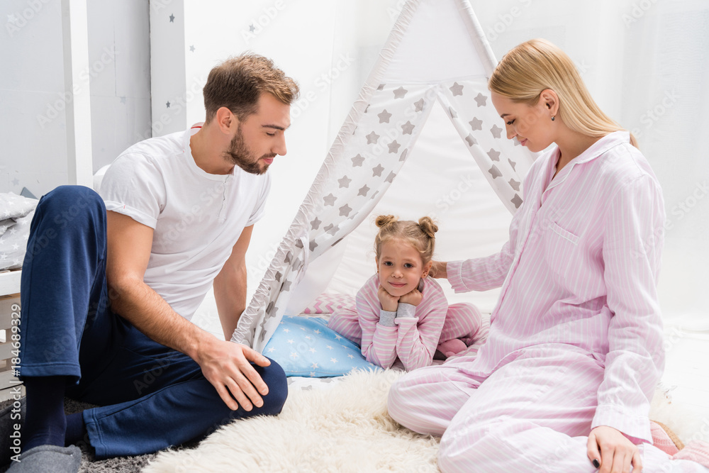 adorable little daughter spending time with parents while relaxing in handcrafted teepee on floor adorable little daughter spending time with parents while relaxing in handcrafted teepee on floor