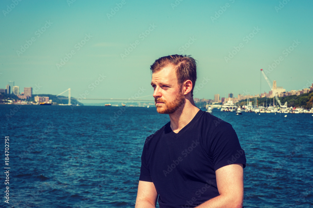 American man traveling in New York. Wearing black v neck T shirt, a young guy with beard, mustache standing by Hudson River, looking forward. Bridge, harbor on background. Filtered effect..