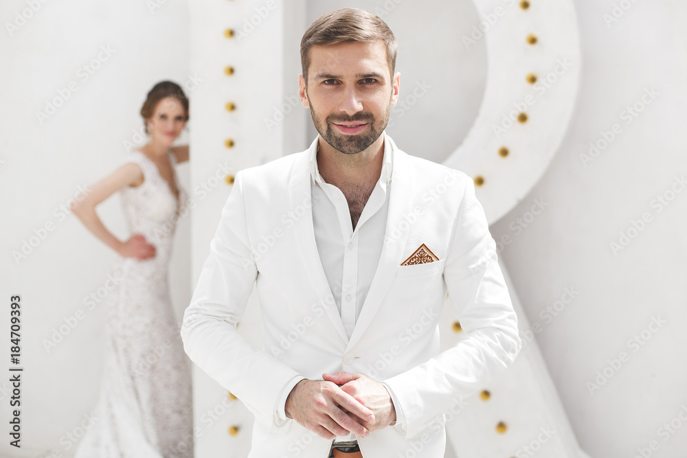 Chic wedding couple groom and bride posing in a white Studio. Happy couple laughing. White dress. White suit. Youth. Wedding. Chandelier. Sofa. The door. Chic wedding couple groom and bride posing in a white Studio. Happy couple laughing. White dress. White suit. Youth. Wedding. Chandelier. Sofa. The door.
