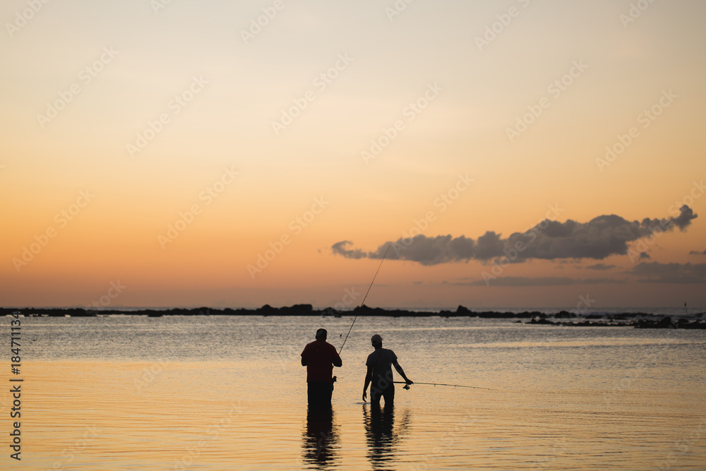 Two men fishing in the ocean from the beach at sunset