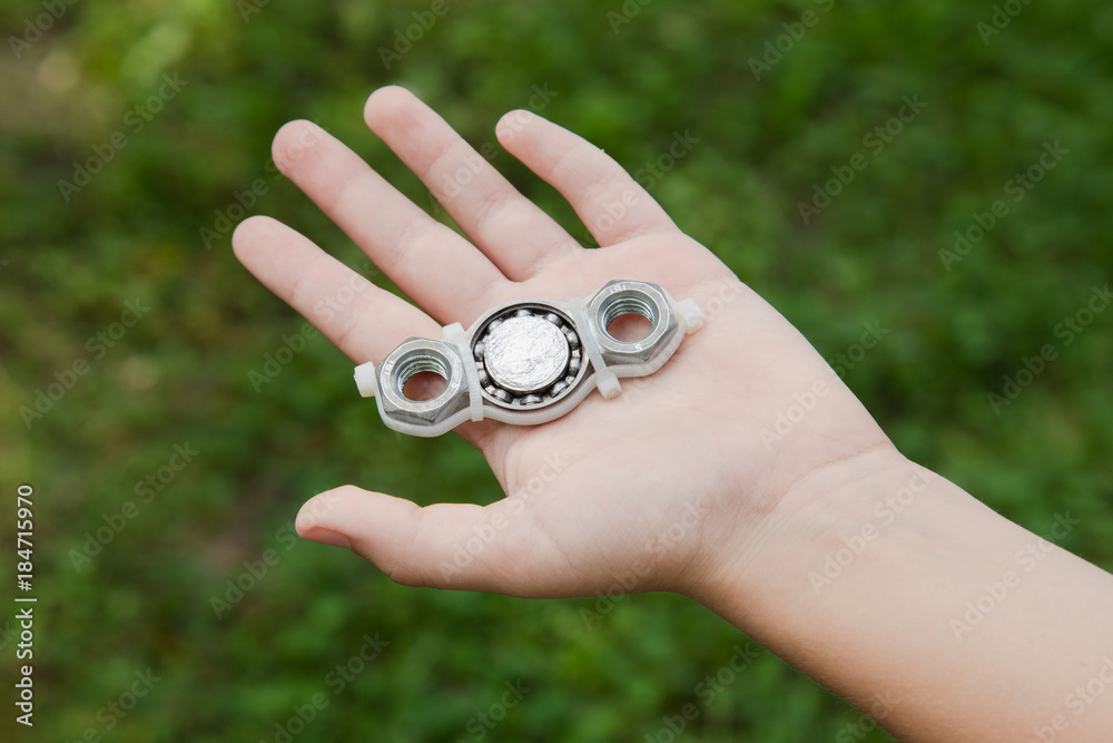 Closeup of hand of white kid playing modern popular hand toy spinner made by himself by using screw-nuts and bearing. Child holding unusual metal spinner isolated at blurry green grass background. Closeup of hand of white kid playing modern popular hand toy spinner made by himself by using screw-nuts and bearing. Child holding unusual metal spinner isolated at blurry green grass background.