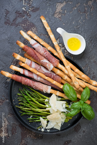 Italian salted breadsticks wrapped with prosciutto and served with fried asparagus and parmesan cheese. Top view on a brown stone background