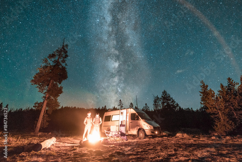 Fototapeta Vanlife couple standing in front of campfire and van under Milky Way night sky