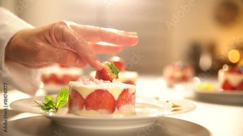 Close-up on the hand of a pastry chef bringing the finishing touch to his strawberry cake by gently placing a strawberry on top.