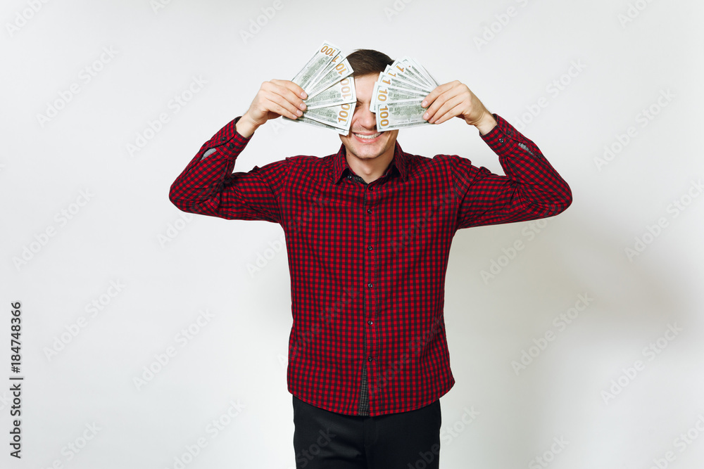 Naklejka premium Handsome cheerful positive satisfied caucasian young happy business man 25-30 years in red shirt hiding behind wad of cash money on white background isolated for advertisement. Winner with dollars.