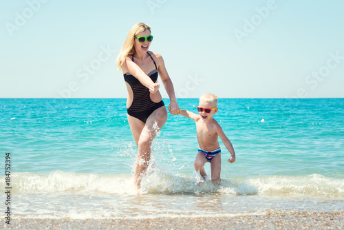 Beautiful young mother and small son holding hands running on the waves on the beach.Fun,family,friendly summer vacation. © Denis