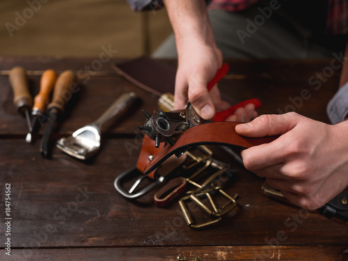 Craftsman holding a hole punch and a piece of leather. On brown wooden table scattered with working tools and accessories. 