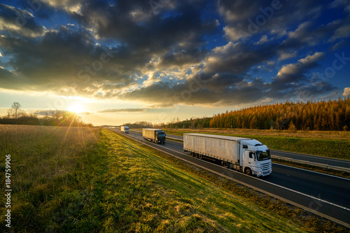 Three trucks driving on a highway in autumn landscape at sunset with dramatic clouds