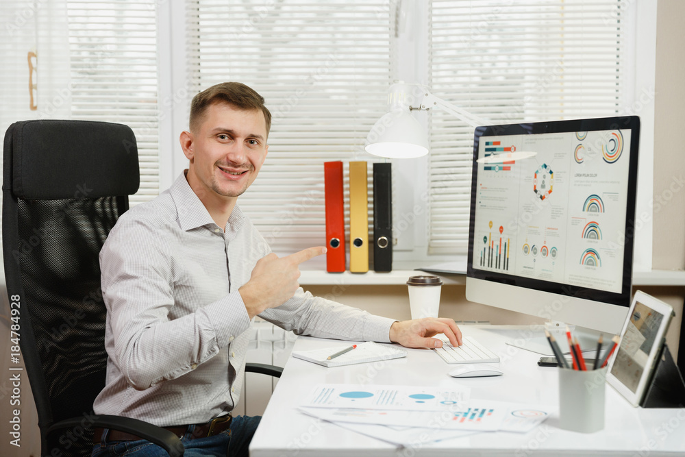 Handsome smiling business man in shirt sitting at the desk, working at computer with modern monitor, lamp, folders, coffee or tea, documents in light office on window background. Manager or worker. Handsome smiling business man in shirt sitting at the desk, working at computer with modern monitor, lamp, folders, coffee or tea, documents in light office on window background. Manager or worker.