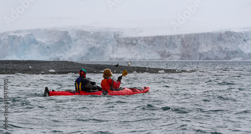 Two kayakers in a red kayak at Yankee Harbor, Antarctica with Gentoo penguin, skua and ice cliffs in background
