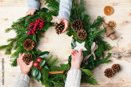 Two small children s hands holding and helping to decorate handmade Christmas wreath made with fir branches  pine cones  berries  baubles on the natural wooden table  top vew  selective focus