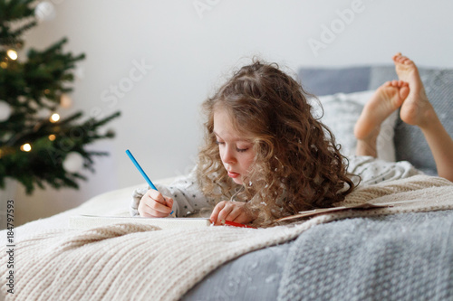 Portrait of adorable curly girl spends free time on bed, makes Christmas card for parents, writes wishes and draws pictures, has concentrated look, being obidient kid. New Year and children concept. photo