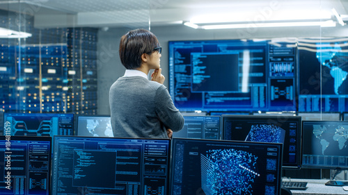 In the System Control Room Technical Operator Stands and Monitors Various Activities Showing on Multiple Displays with Graphics. Administrator Monitors Work of  Artificial Intelligence. photo
