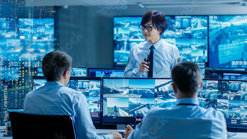 In the Security Control Room Chief Surveillance Officer Briefs Two of His Subordinates. Multiple Screens Show that They Guard Object of Big Importance.