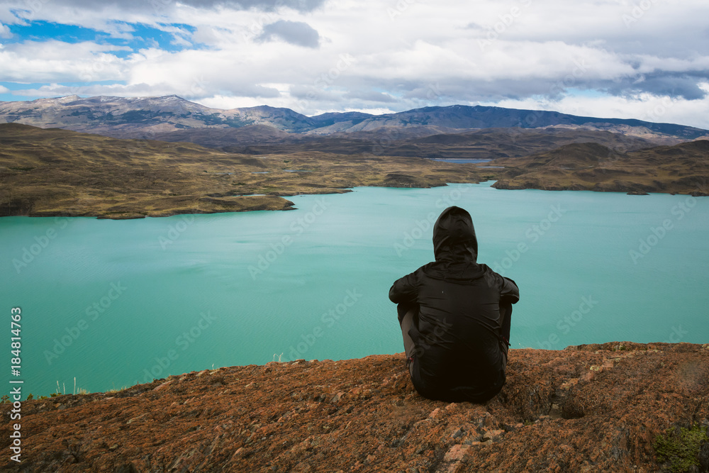 Young man is sitting above the turquoise colored lake  in Torres Del Paine National Park in Patagonia, Chile