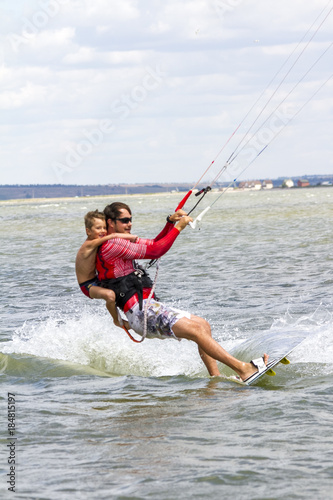 Dad and son are kitesurfing on the sea. The son clings to Papa's back
