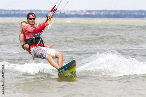 Dad and son are kitesurfing on the sea. The son clings to Papa's back