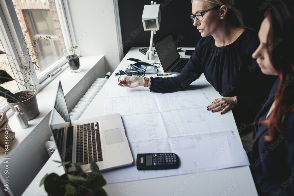 Female engineers discussing over blueprint on desk at home office Female engineers discussing over blueprint on desk at home office