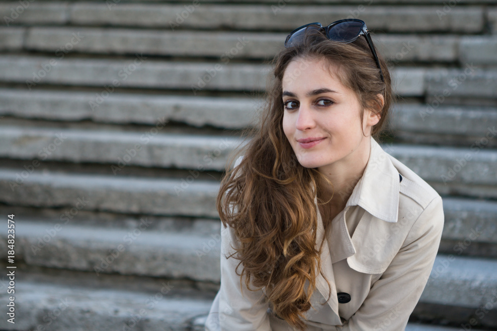 Young attractive woman wearing beige jacket and sunglasses smiling over stairs. Beautiful long curly hair. Young attractive woman wearing beige jacket and sunglasses smiling over stairs. Beautiful long curly hair.