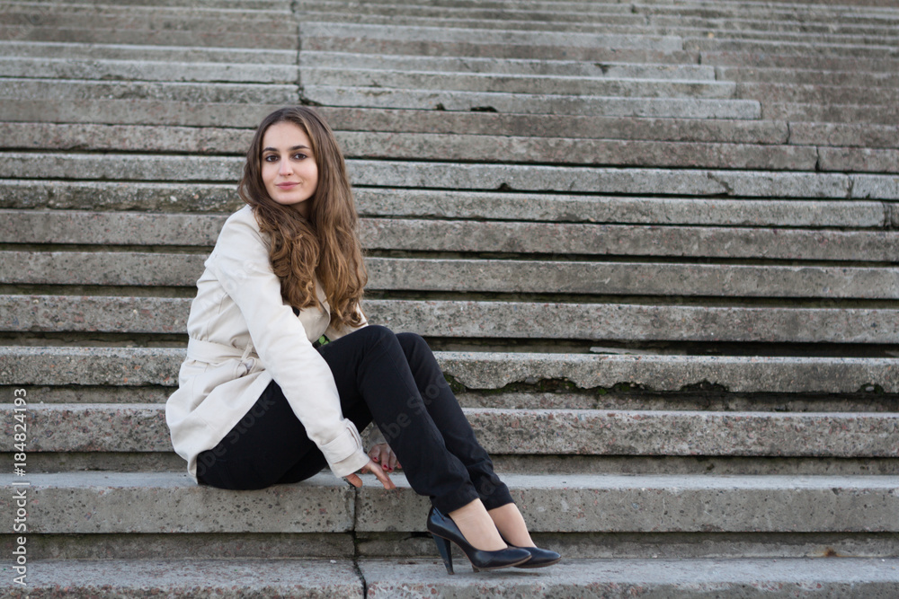 Young beautiful woman wearing beige jacket sitting on concrete stairs Young beautiful woman wearing beige jacket sitting on concrete stairs