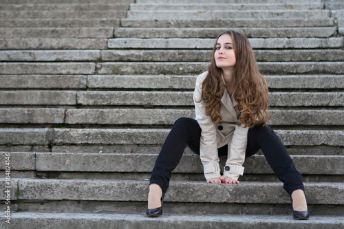 Young beautiful woman wearing beige jacket sitting on concrete stairway and looking aside