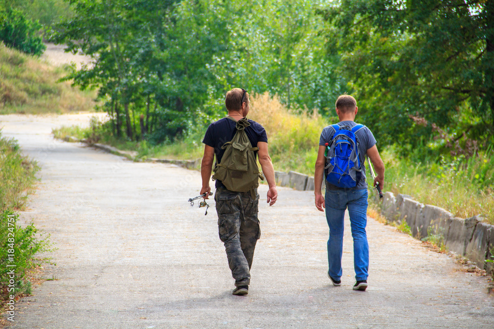 Two fishermen go fishing with fishing rods along the asphalt path through the forest. Two fishermen go fishing with fishing rods along the asphalt path through the forest.