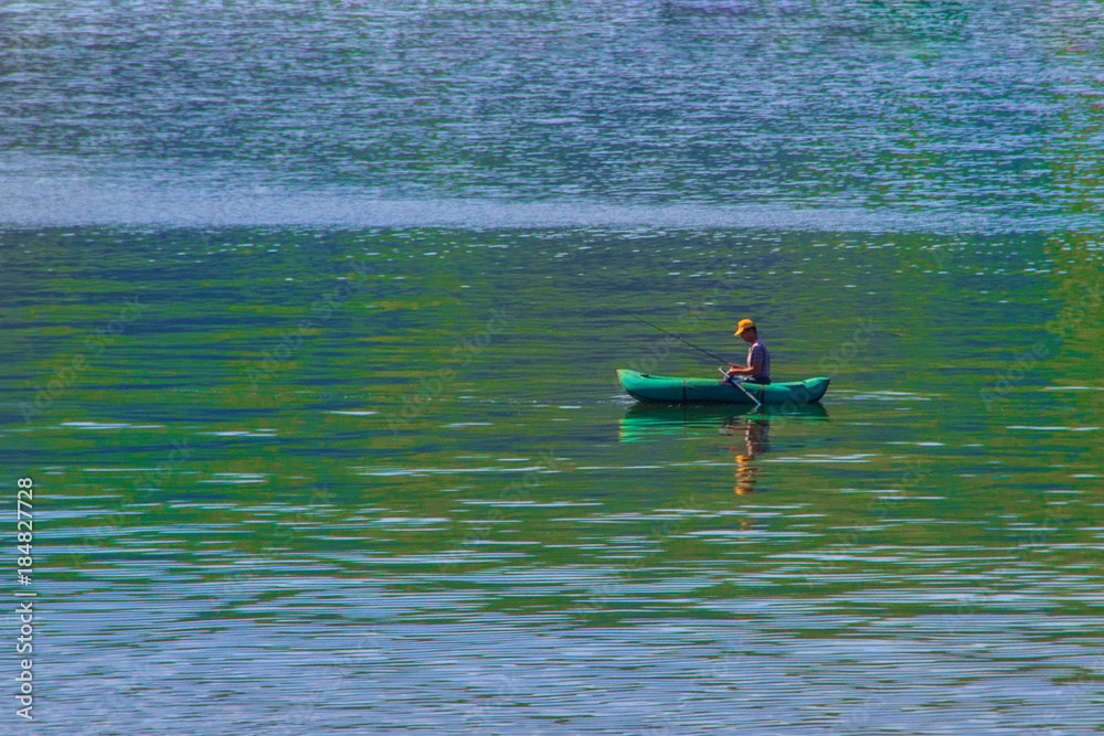 A fisherman with a fishing rod swims along the river in a green inflatable rowing boat to fish.