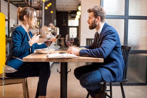 Fototapeta Naklejka Na Ścianę i Meble -  Caucasian business couple dressed strictly in the suits having discussion sitting together at the restaurant during the dinner