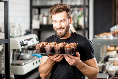 Portrait of a handsome bearded confectioner in black t-shirt and apron holding mafins at the cafe photo