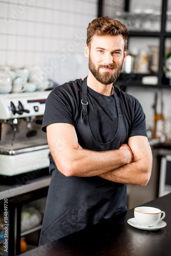 Portrait of a handsome barista in black t-shirt and apron standing at the bar of the modern cafe