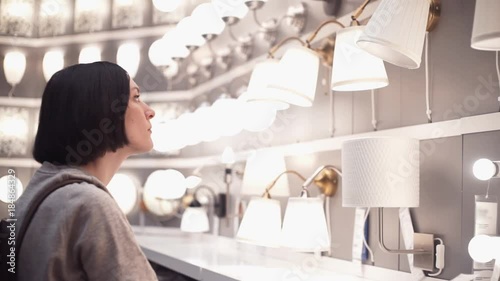 Customer chooses bedroom goods in hypermarket. Young woman examining lamp