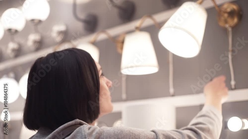 Purchaser chooses bedroom goods in hypermarket. Young woman examining lamp