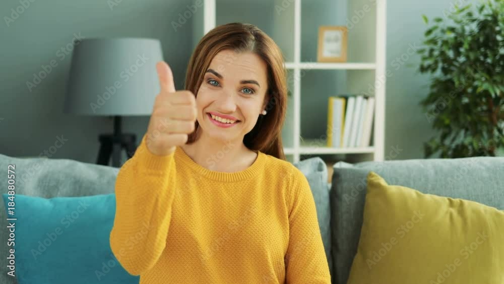 Cheerful pretty woman sitting on the grey sofa with pillows and giving her thumb up in the home room. Portrait shot. Indoor Cheerful pretty woman sitting on the grey sofa with pillows and giving her thumb up in the home room. Portrait shot. Indoor