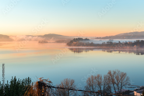 Lake Varese  Gavirate  province of Varese  north Italy. Picturesque sunrise with fog. Below is Gavirate visible  in the background the village of Biandronno and the islet Virginia  UNESCO site