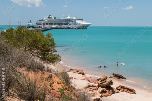 Couple swimming with dogs with modern cruise ship tied up to jetty surrounded by a turquoise sea in the background at Broome in Western Australia.