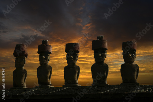 Fünf Moai Statuen mit Kopfbedeckung vor einem dramatischen Wolkenhimmel am Abend.
