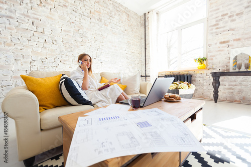 Female freelancer in a white coat clothing working remotly from her dining table in the morning. Houses on a sofa on a white brick wall background. Architect, blueprints. talking on the phone. photo