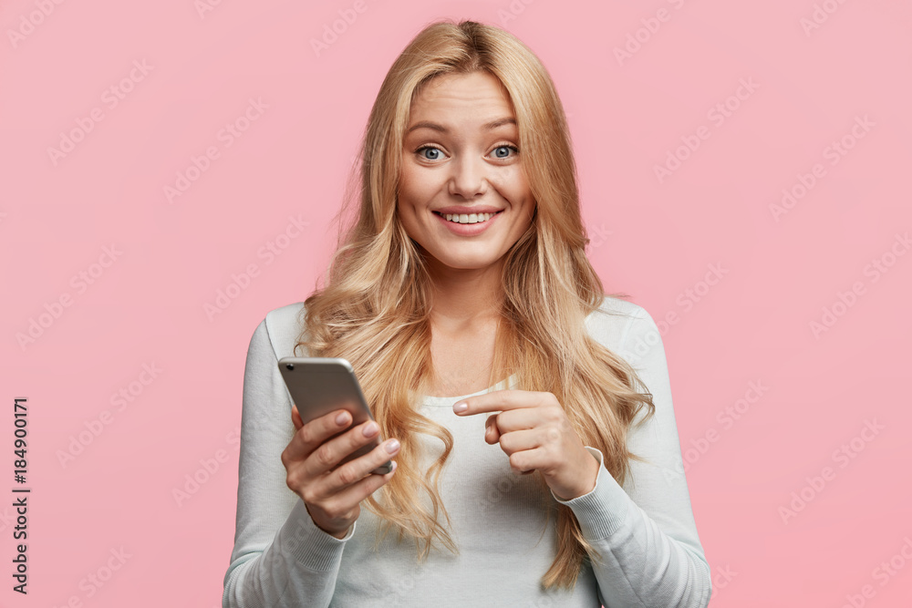Φωτογραφία Cheerful attractive female model checks in network on smart phone or read good news, indicates into screen, wants to present some information, isolated over pink background