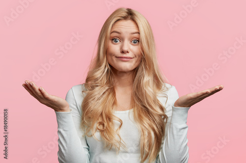 Pretty blonde female with blue eyes and blonde hair, shrugs shoulders, being doubtful about making serious decision, feels uncertain, isolated over pink background. Hesitation and confusion.