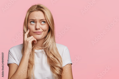 Indoor shot of thoughtful pretty woman has long light hair, looks aside with pensive expressions, plans something on coming weekends, poses against blank copy pink space in studio. Dreamful female
