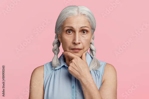 Mature confident serious woman with grey pigtails, wears fashionable blouse, watches news attentively, keeps hand on chin, isolated over pink background. Advanced age. Wrinkled female pensioner photo