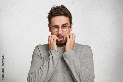 Scarying bearded male model with beard and mustache looks nervously into camera, bites nails with fright, isolated over white concrete background. Man has digusting expression. Negative emotions