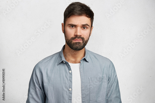 Portrait of serious bearded male wears fashionable denim shirt, hears attentively something important, isolated over white concrete wall. Young male student with thick beard and mustache indoor photo