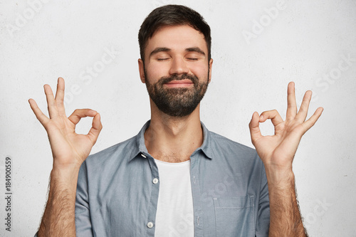 Positive bearded male model wears denim shirt, makes ok gesture and closes eyes, has happy expression, isolated over white background. Optimistic male expresses his approval. Body language concept
