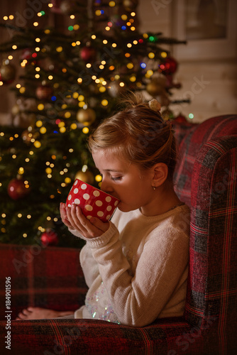 Girl drinking cocoa from a mug by a christmas tree