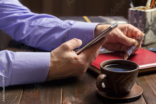 Home office workplace, male hands hold a smartphone and credit card, notepad, cup of black coffee on dark wood working table, business, finance concept