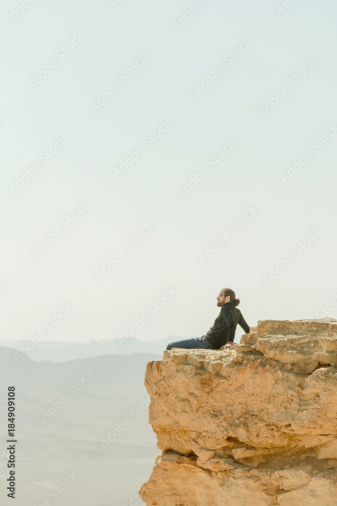 Alone charismatic young man sitting on the cliff in desert