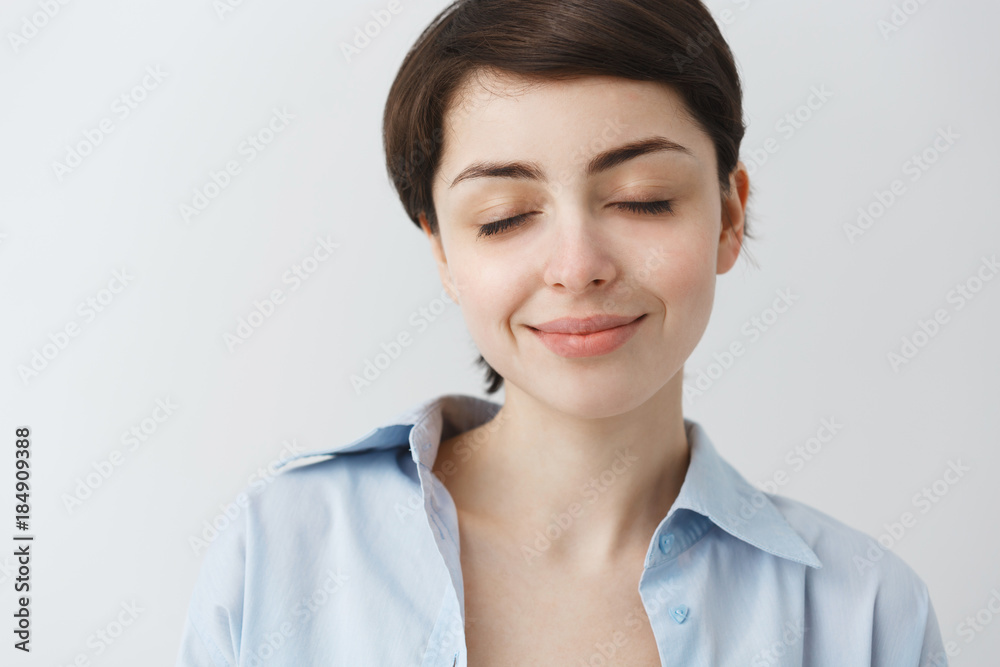 Close up of attractive young girl with dark short hair in blue shirt smiling gently with closed eyes having relaxed and calm look. Close up of attractive young girl with dark short hair in blue shirt smiling gently with closed eyes having relaxed and calm look.